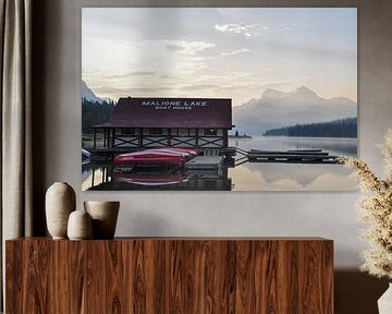 A serene lake in Canada, surrounded by majestic mountains and lush green trees, under a blue sky during sunrise, with dew on the surface of the water and a boathouse in the foreground. The calm water reflects the landscape, creating a sense of symmetry and balance. The landscape exudes peace and serenity. by UMA Digital NL