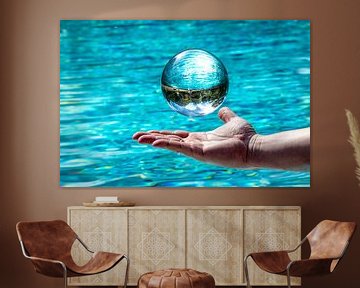 Glass ball hovering over a hand in front of water background with mirrored cloudy sky and trees by Hans-Jürgen Janda