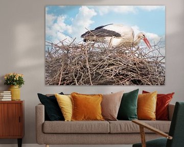 A stork stands in the nest, twig in its beak. Blue sky with white clouds in the background.  Greetin